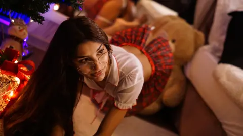 Young woman in glasses and red plaid outfit relaxing near a teddy bear in a cozy, festive holiday setting.