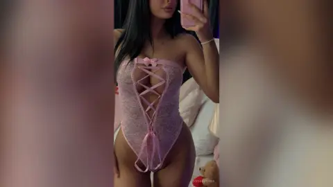 Woman in a pink lace corset bodysuit posing in a bedroom setting with a romantic mood.