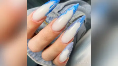 Close-up of long stiletto nails with white and blue marble swirl nail art design on a hand wearing denim.