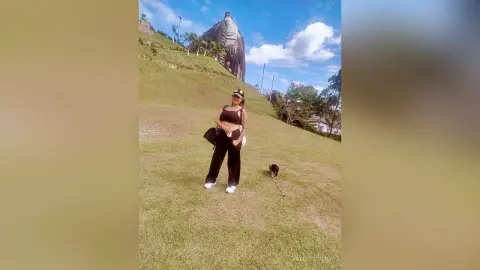 Smiling woman in black tank top and pants walking her dog on a grassy hill near a large stone monument.