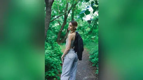 Young woman in a striped crop top and light denim jeans standing in a lush green forest setting.