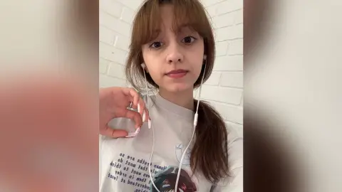 Young woman with brown hair wearing a white graphic t-shirt and wired earbuds in a bright room.