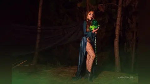 AmberNash — Mystical woman in a dark blue flowing gown holding a glowing green orb in a spooky, dark forest setting.