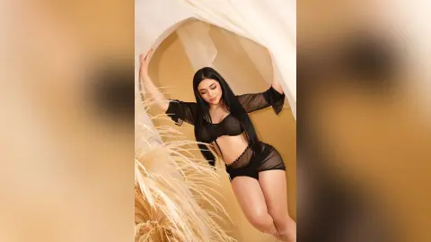 IvyStuart poses with her arms up, wearing a black sheer mesh top and matching black bottoms against a tan background.