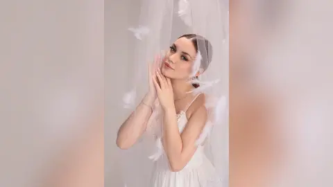 Elegant woman in a white lace dress posing dreamily behind sheer white fabric in a soft, ethereal setting.