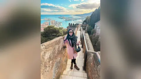 Aleja stands on a stone walkway overlooking a coastal city and ocean, wearing a pink coat, black scarf, and sunglasses.