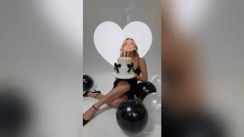 SerenaStar wears a black dress while holding a small cake with lit candles, surrounded by black balloons against a heart-shaped background.