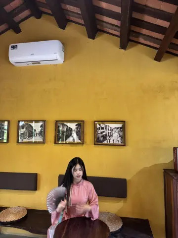 Young woman in a pink blouse holding a traditional fan in a rustic cafe with yellow walls.