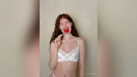 Redheaded woman in white lace lingerie eating a red lollipop against a neutral background.