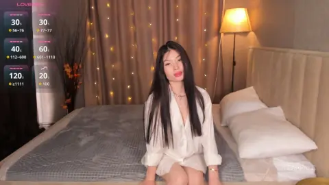 Young woman in a white button-down shirt sitting on a bed in a cozy, dimly lit bedroom with fairy lights.