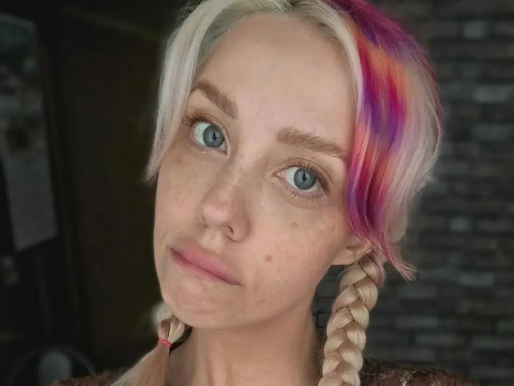 Close-up portrait of a blonde woman with pink hair highlights and blue eyes looking thoughtfully at the camera.