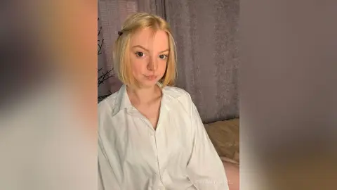 Blonde woman in a white button-down shirt sitting in a cozy room with soft lighting.