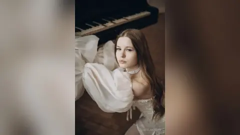 StelaBrown poses in a white puff-sleeve dress and pearl choker while standing next to a black piano.