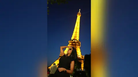 Dolce poses in front of the lit Eiffel Tower at night wearing a black top and sunglasses.