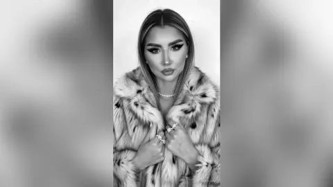 Glamorous woman in a luxury faux fur coat and pearl necklace posing with elegant makeup in a black and white portrait.