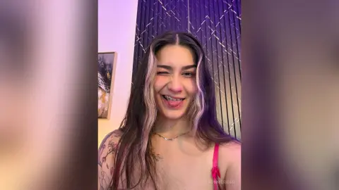 BonnieRider winks and sticks out her tongue, showing her teeth and tongue while wearing a pink top and a silver necklace.