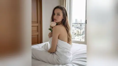 Elegant woman in white loungewear sitting on a bed with a rose in a bright, serene bedroom setting.