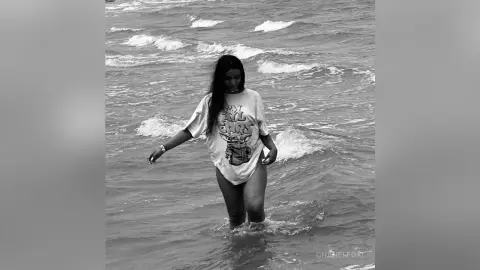 Young woman in a graphic t-shirt standing in ocean waves during a moody black and white beach photoshoot.