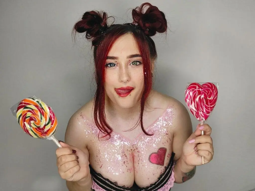 Redheaded woman with glittery skin holding colorful heart-shaped lollipops in a playful studio setting.
