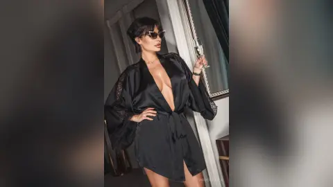 Stylish woman in a black satin robe and sunglasses posing elegantly in a modern luxury interior.