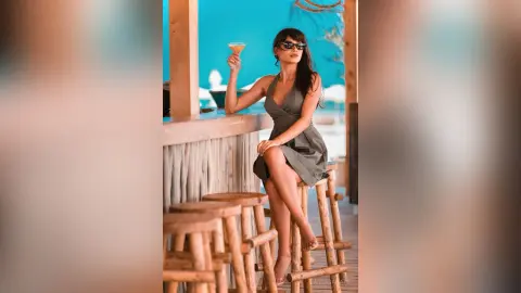 Stylish woman in a grey dress holding a cocktail at a tropical beach bar during sunset.