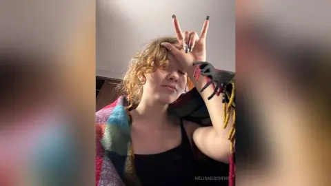 Smiling young woman with curly hair wearing a black tank top and colorful patterned shawl in a cozy indoor setting.