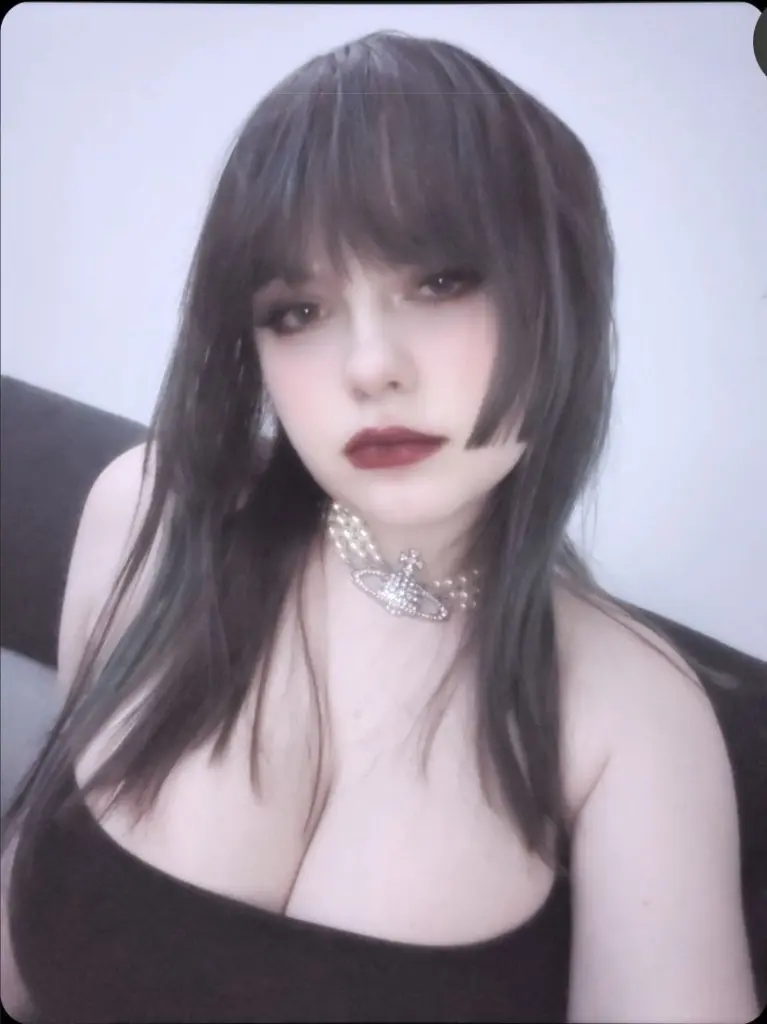 LoveluKristi poses with her hand near her mouth, showing her long dark hair and black tank top in a dimly lit room.