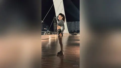 Stylish woman in a grey crop top and black shorts posing on a wooden pier at night under city lights.