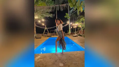 LaurentBelluci stands at the edge of a bright blue swimming pool wearing a white top and a long, patterned skirt.