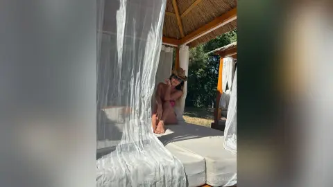 TiffanyAddams sits on a white bed under a thatched cabana, wearing sunglasses and a pink bikini while posing outdoors.