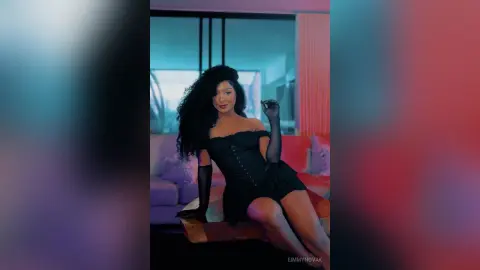 Glamorous woman with curly hair in a black strapless mini dress sitting in a moody, dimly lit modern living room.