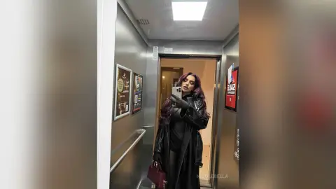 NicoleBella poses in a mirror selfie wearing a black leather coat and black top while standing in an elevator.