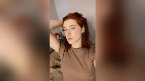 KeithaHall poses with her red hair and a brown t-shirt, resting her hand behind her head against a plain wall.