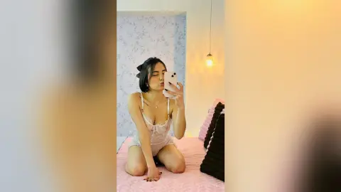 Young woman in white lace lingerie sitting on a bed in a bright, cozy bedroom taking a selfie.