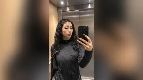Young woman with curly hair in a black zip-up jacket taking a moody selfie in an elevator mirror.