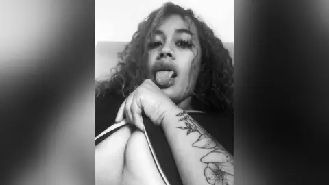 Black woman with curly hair and arm tattoos sticking her tongue out in a playful black and white portrait.