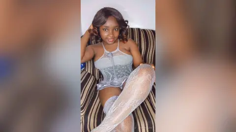 Smiling young woman in a grey lace bodysuit and white fishnet tights sitting on a striped armchair.