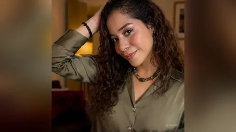 Smiling woman with curly hair wearing a green satin blouse and beaded necklace in warm indoor lighting.