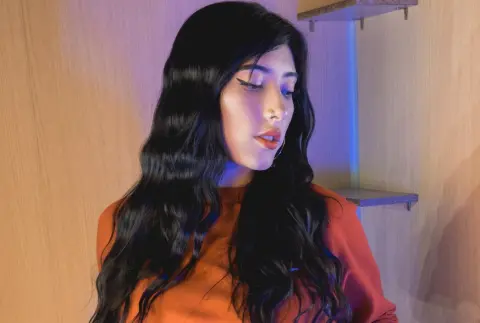 Young woman with long wavy black hair wearing an orange top in moody blue and warm ambient lighting.