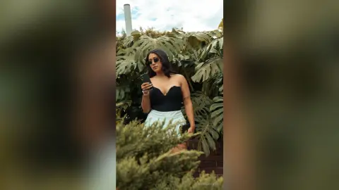 Stylish woman in a black bodysuit and white skirt taking a selfie amidst lush tropical monstera leaves.