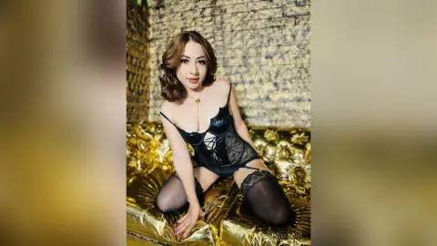 Glamorous woman in black lace lingerie and stockings sitting on a gold sequin sofa against a textured wall.