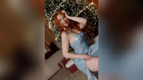 Smiling redhead woman in a sparkling blue sequin dress posing by a glowing Christmas tree.