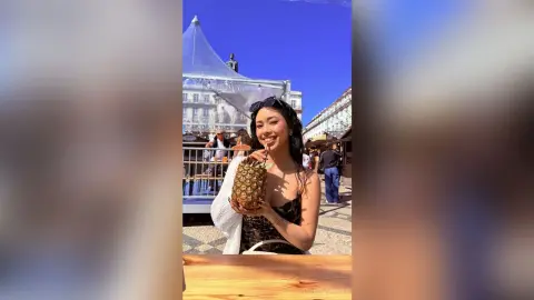 Smiling woman in a black dress holding a pineapple drink at an outdoor sunny cafe terrace.