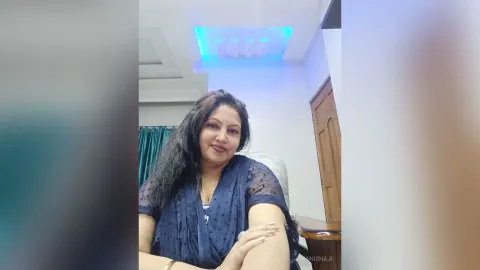 ManishaJi — ManishaJi sits in a chair wearing a navy blue sheer blouse with her long dark hair flowing over her shoulder.