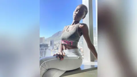 Confident woman in grey sports bra and leggings posing by a bright window overlooking a city skyline.