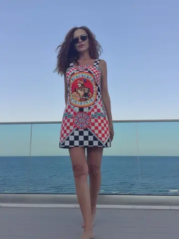 Woman in a colorful graphic print sundress standing on a sunny seaside balcony overlooking the ocean.