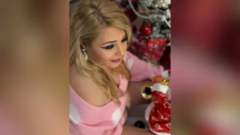 Blonde woman in a pink sweater smiling near a festive Christmas tree in a cozy holiday setting.