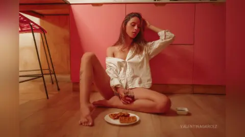 ValentinaGarcez sits on a wooden floor in an open white shirt, posing with one hand in her hair near a plate of waffles.