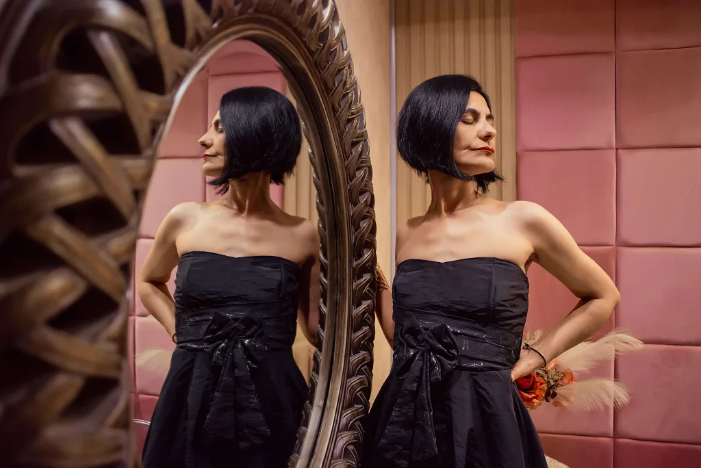 KalystaSwiss poses with one hand behind her head, wearing a black strapless dress against a round mirror and warm indoor background.