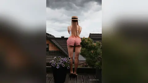 AdelinaKael stands outdoors on a balcony wearing red checkered shorts and a white top, showing her long blonde hair and bare legs against a cloudy sky.